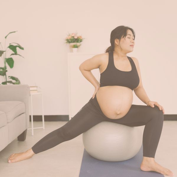 Pregnant woman stretching on a stability ball in a living room, practicing prenatal mobility or pelvic opening exercises.